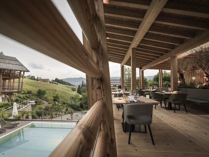Genussreisen und Wellness in Völs am Schlern Überdachte Terrasse mit Tischen und Stühlen, Blick auf Pool und grüne Hügel