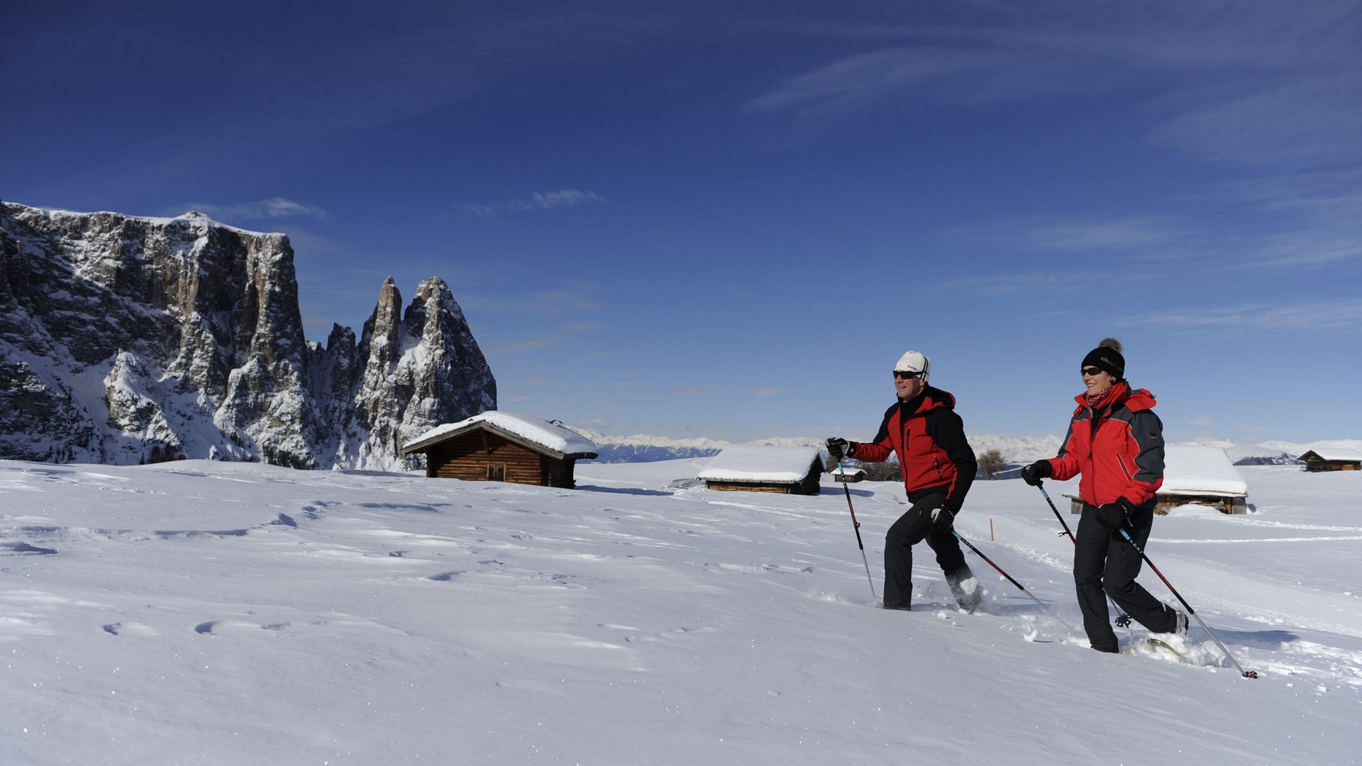 Due persone che fanno escursioni con racchette da neve davanti a baite e montagne innevate