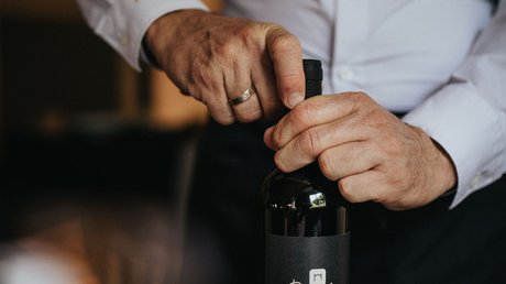 Immagini che emozionano: appartamenti Presulis a Fiè Persona che apre una bottiglia di vino con etichetta nera su tavolo apparecchiato