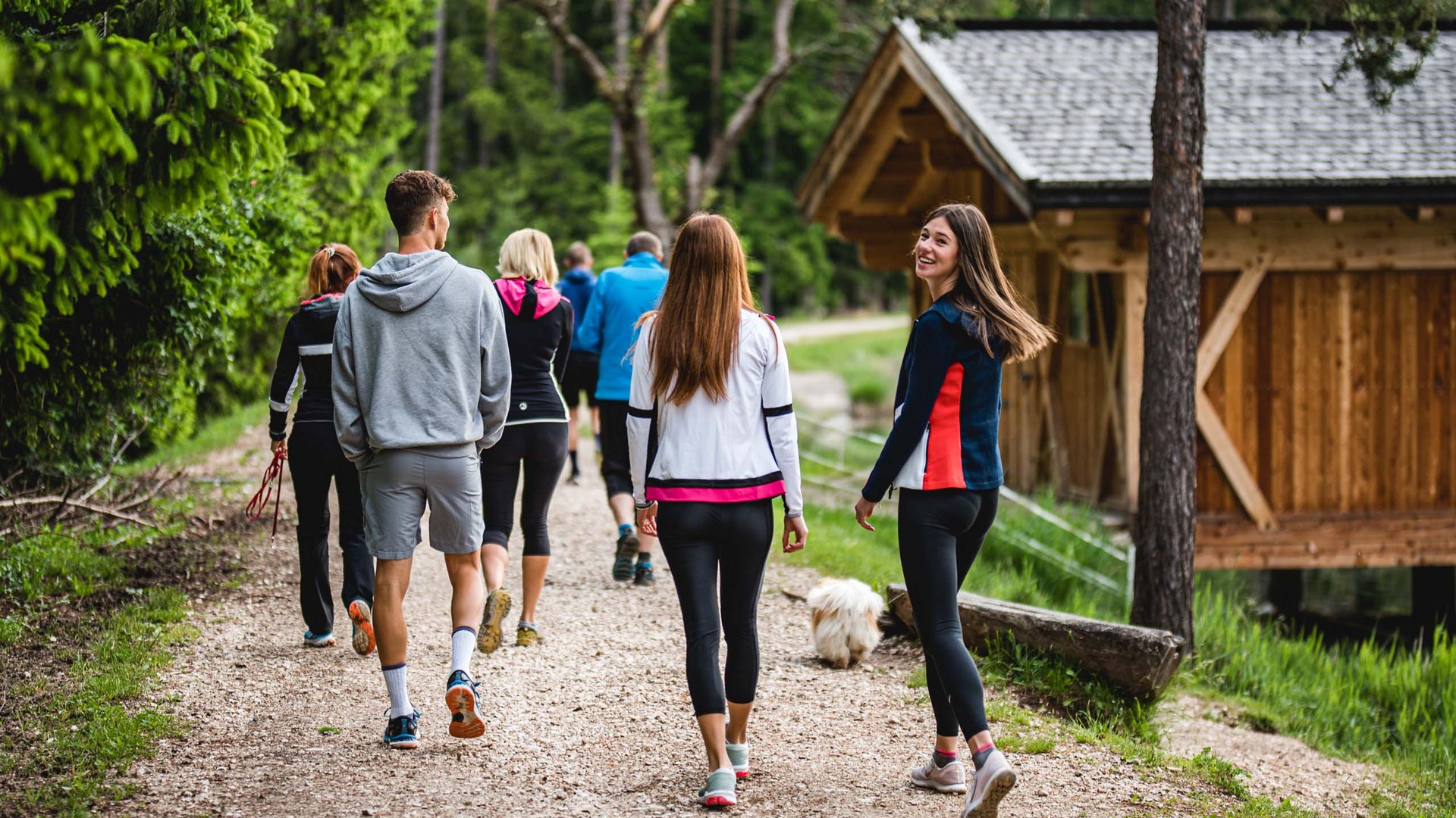 Seiser Alm Balance: active & healthy throughout spring Group walking on forest path near wooden cabin, one woman smiling back