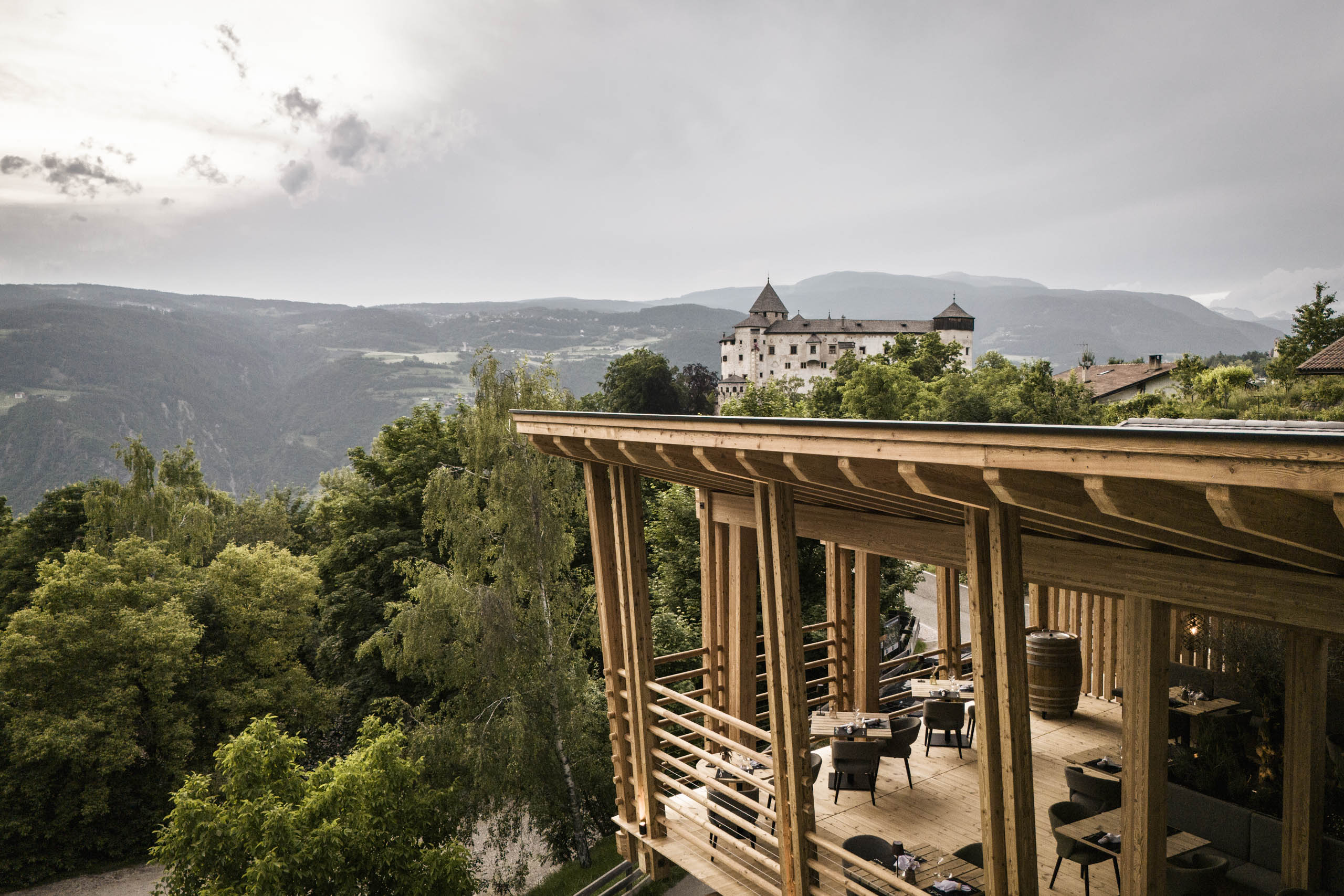 Alloggi all’Alpe di Siusi | Appartamenti a Fiè allo Sciliar Terrazza in legno con vista su un castello e montagne boschive sullo sfondo